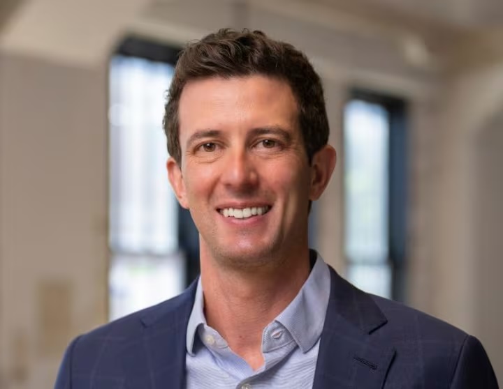 A man with short brown hair wearing a blue blazer and light blue shirt smiles in a brightly lit indoor setting with blurred windows in the background.