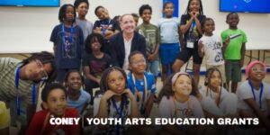 A group of smiling children and one adult pose together in a classroom. Text reads “Coney Youth Arts Education Grants.”.