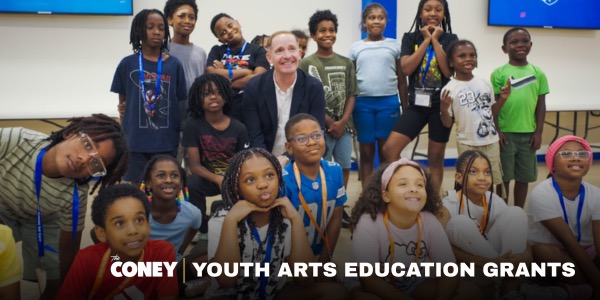 A group of smiling children and one adult pose together in a classroom. Text reads “Coney Youth Arts Education Grants.”.