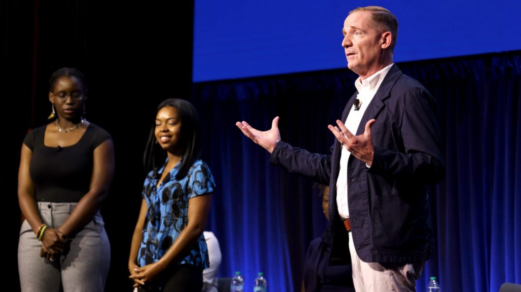 A man speaks on stage with his hands raised, while two women stand nearby listening. A blue screen and dark curtains are in the background.